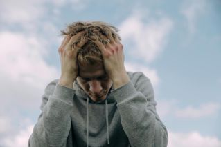 Young man in gray hoodie holds head in frustration, set against cloudy sky.