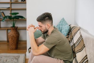 A young man with clasped hands, sitting thoughtfully on a sofa in a cozy living room.