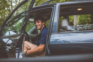 A cheerful man sits inside a car with the door open, enjoying a sunny day outdoors.